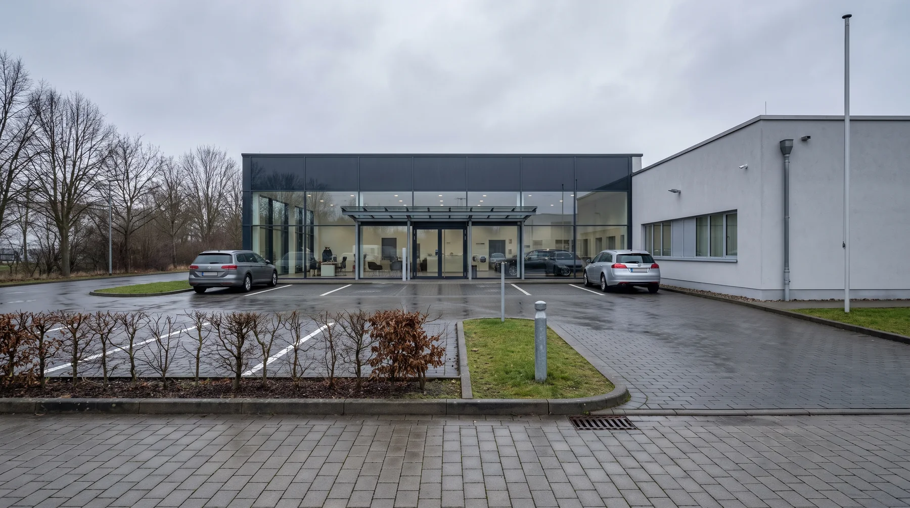 Derselbe Vorplatz von außen: Pflasterfläche mit Stellplätzen, Hecke, Rasenstreifen und die Glasfront des Eingangs